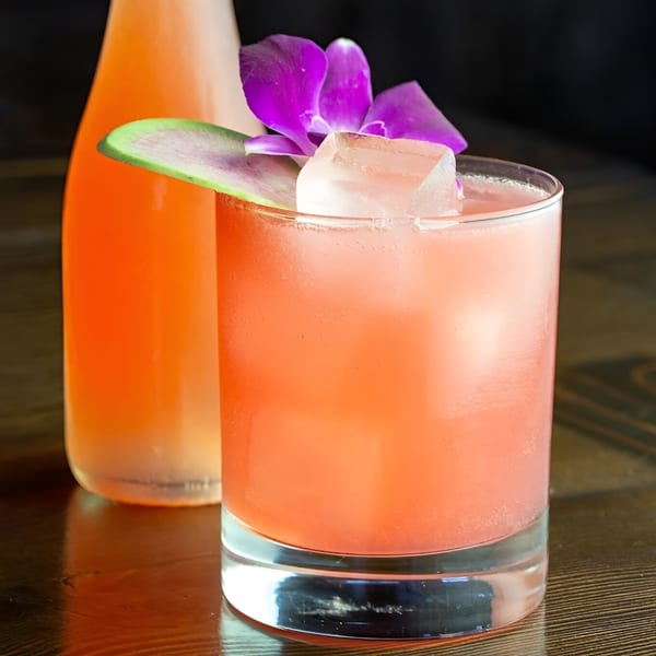 A pink cocktail with ice, a purple orchid, and a watermelon slice, next to a bottle.