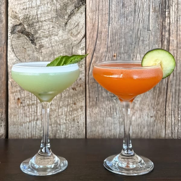 Two cocktails, one green with basil and one orange with cucumber, against a rustic wooden wall.