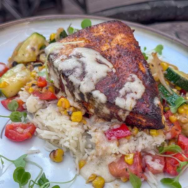 Seared fish with melted cheese, rice, corn, zucchini, and diced tomatoes on a white plate.