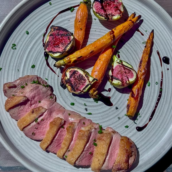 Sliced duck breast with crispy skin, roasted carrots, grilled figs, and balsamic glaze on a white plate.
