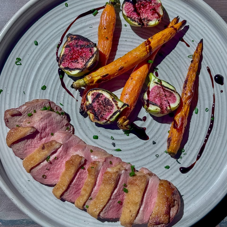 Sliced duck breast with crispy skin, roasted carrots, grilled figs, and balsamic glaze on a white plate.