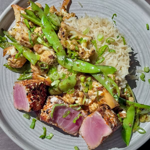 Seared tuna with sesame crust, rice, roasted cauliflower, and snap peas on a gray plate.
