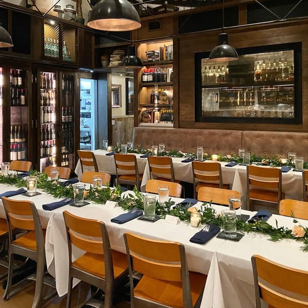 Long dining tables set with white tablecloths, green garlands, and wooden chairs in a dimly lit Craft House.