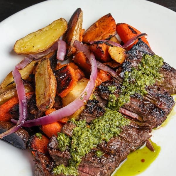 Grilled steak with green chimichurri sauce, roasted potatoes, carrots, and red onions on a white plate.