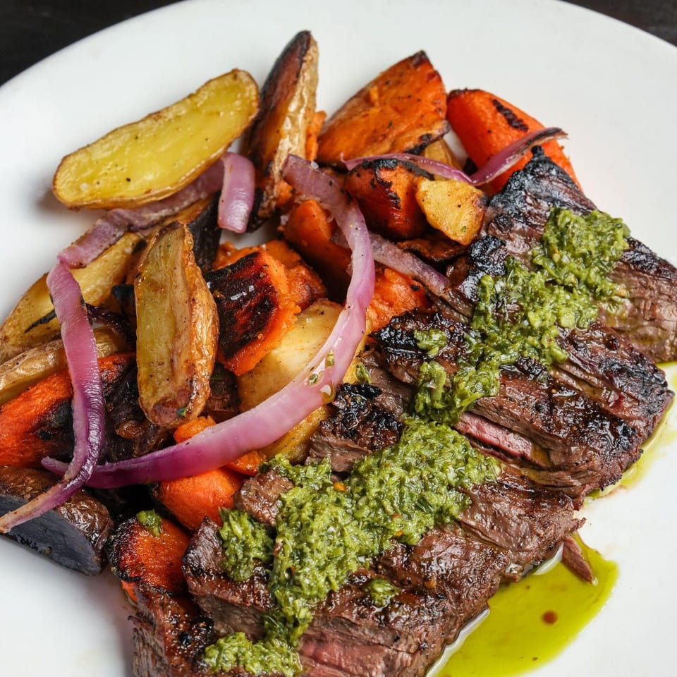 Grilled steak with green chimichurri sauce, roasted potatoes, carrots, and red onions on a white plate.